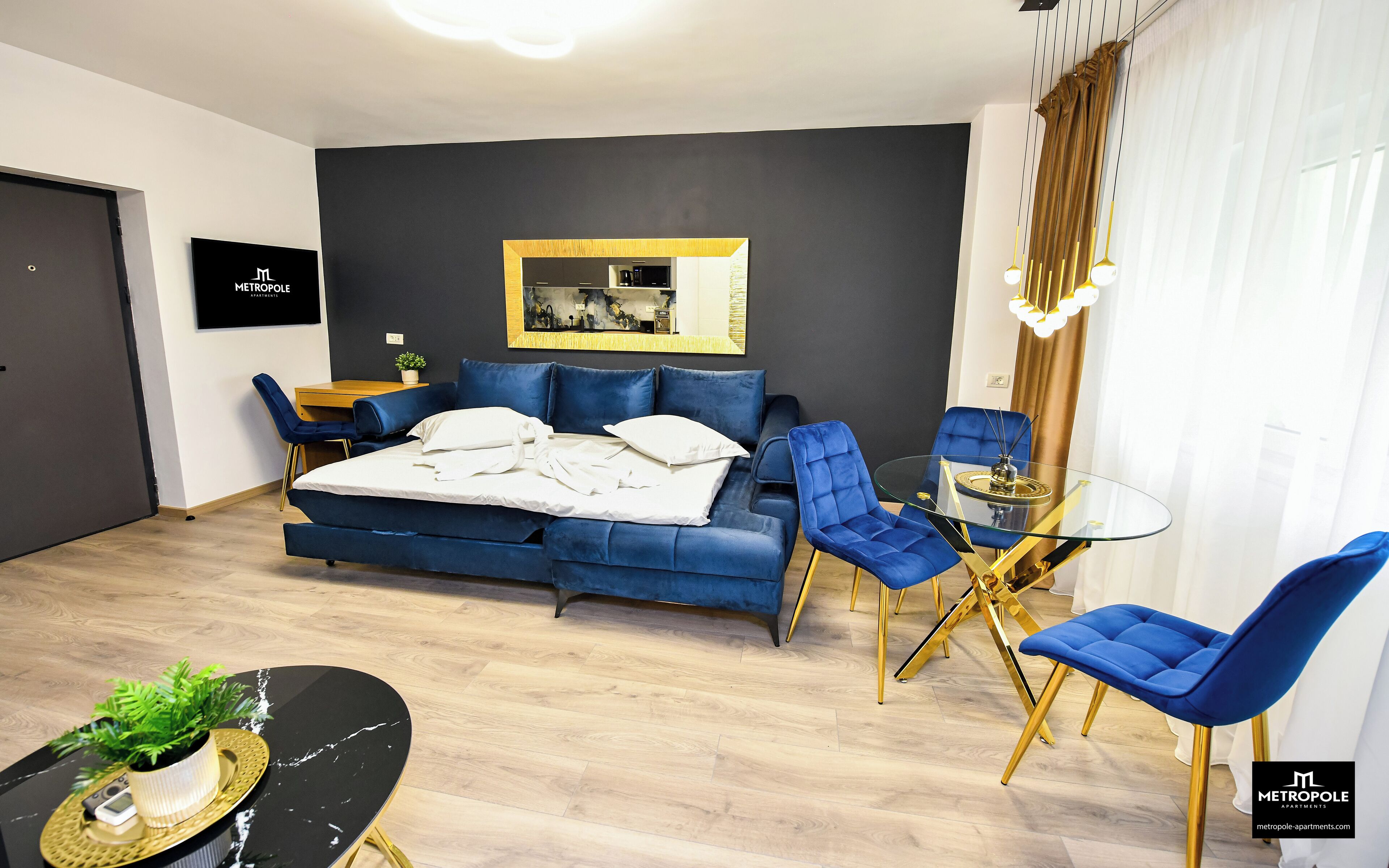 A modern bedroom with a blue sofa bed, a glass dining table with blue chairs, a small desk, and a potted plant.
