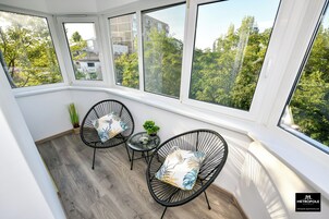 Una habitación luminosa con ventanas amplias, dos sillas de alambre negras con cojines estampados, una mesita redonda y una planta en maceta.
