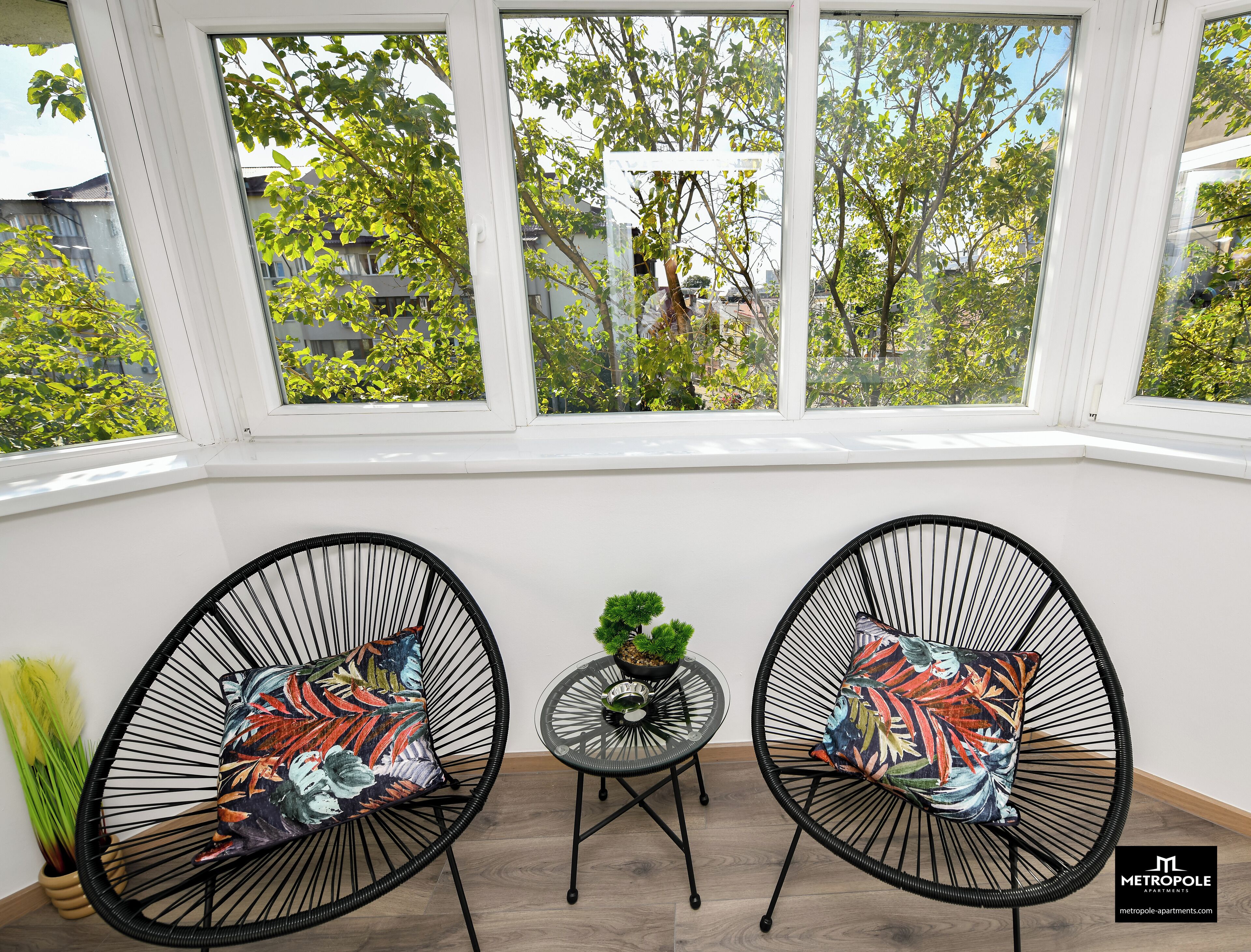 Una habitación con dos sillas de alambre negro y cojines de colores, una pequeña mesa redonda con una planta verde, y vistas a árboles a través de amplias ventanas.