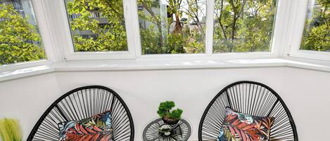 Una habitación con dos sillas de alambre negro y cojines de colores, una pequeña mesa redonda con una planta verde, y vistas a árboles a través de amplias ventanas.