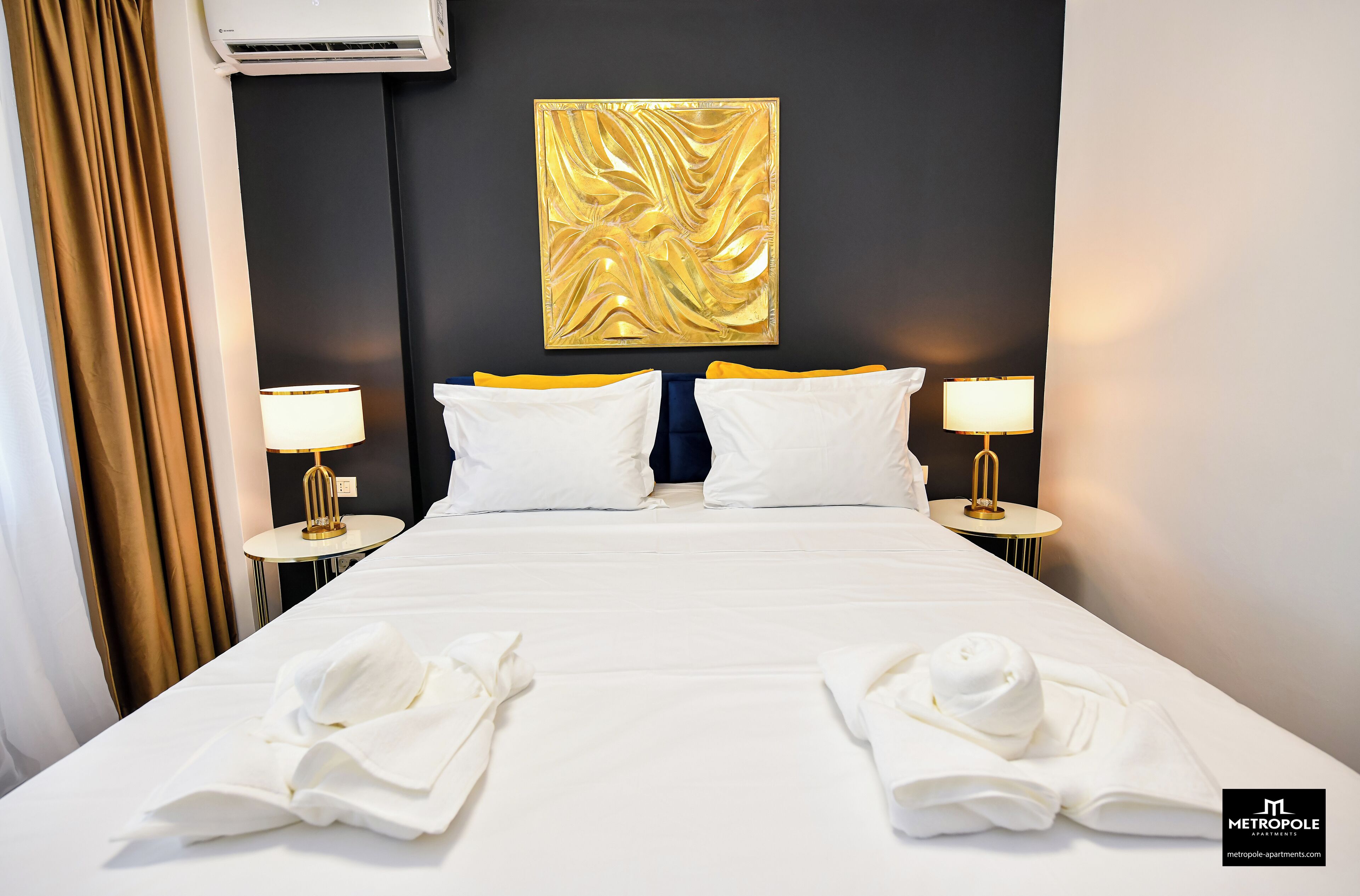 Une chambre d’hôtel avec un grand lit, deux tables de chevet avec des lampes, une œuvre d’art abstraite dorée accrochée au mur et un mur d’accent de couleur sombre.