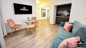 A modern living room with a teal sofa, pink chairs, a glass dining table, and a kitchen area with black cabinets.