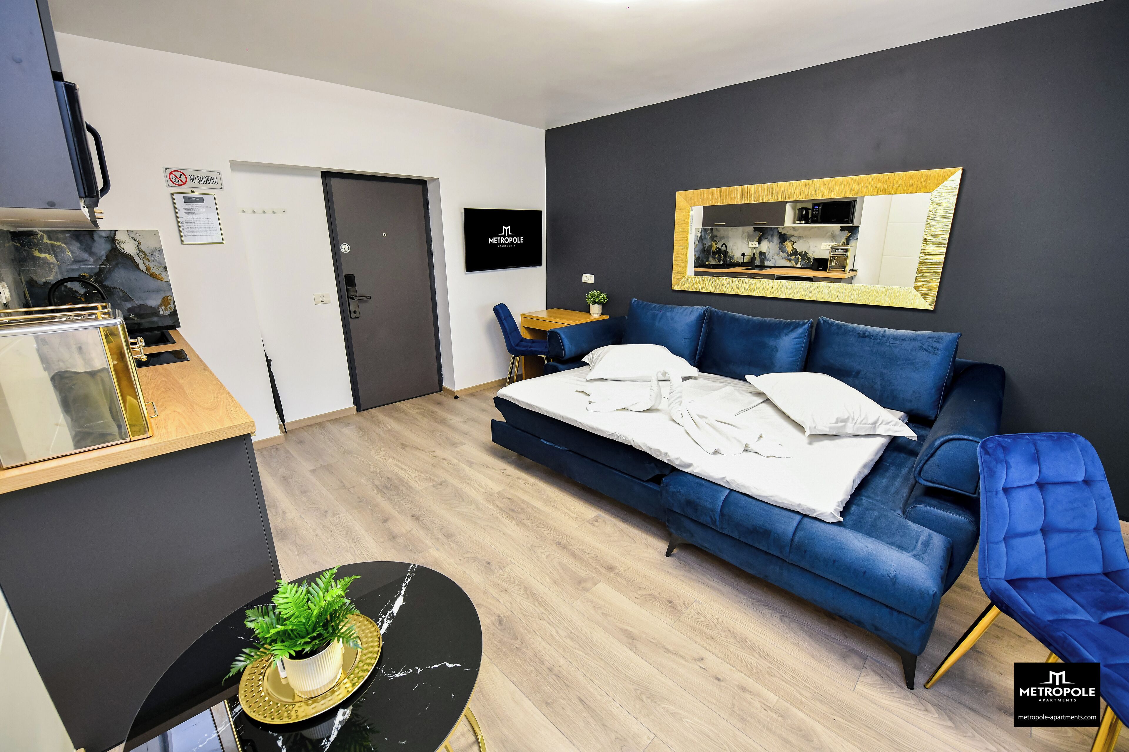 A modern hotel room with a blue sofa, a dining table with a plant, a kitchen area, and a 'No Smoking' sign.