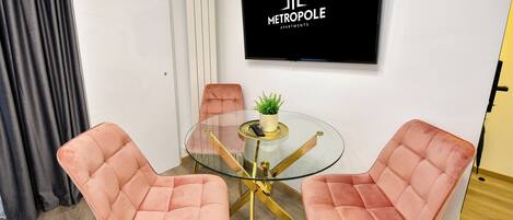 Uma sala de estar moderna com uma mesa de vidro, duas cadeiras cor-de-rosa, um vaso de planta e uma TV exibindo o logotipo de 'Metropole Apartments'.
