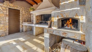 Une cheminée extérieure en pierre, avec un feu qui crépite, entourée de murs en pierre et surmontée d’un plafond en bois.