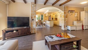 Un salon avec un téléviseur, une table basse sur laquelle on peut voir des fruits et des livres, et une cuisine en arrière-plan.