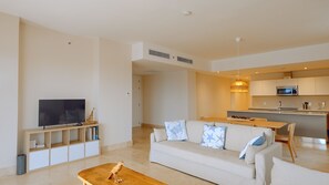 Uma sala de estar moderna com televisão de tela plana, mesa de centro de madeira, sofá bege com almofadas estampadas e um espaço de cozinha com micro-ondas e luminária pendente.