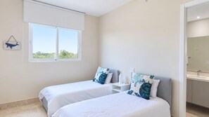 Un dormitorio con dos camas, una ventana, una mesita de noche y un baño visible a través de una puerta abierta.