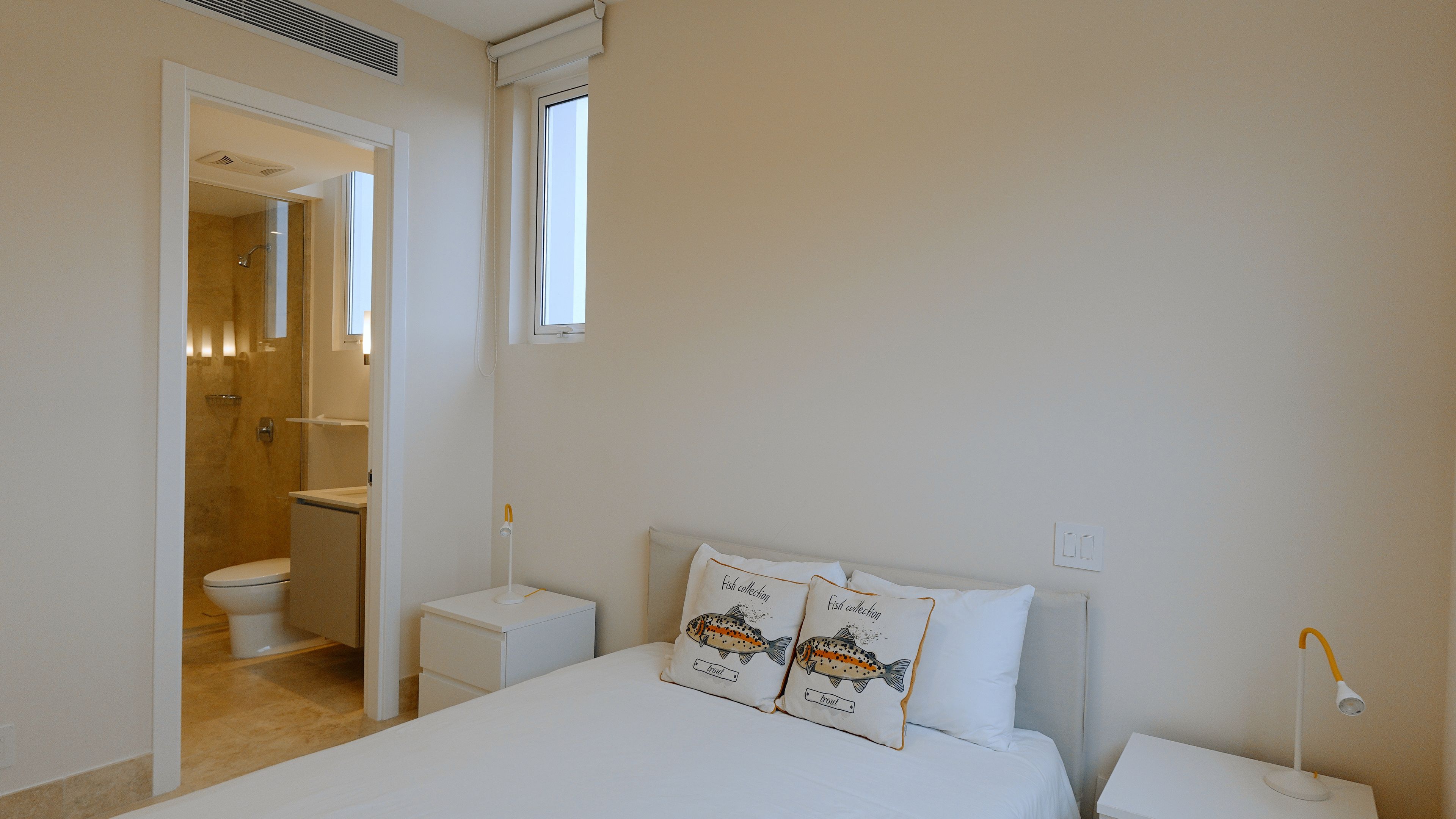 A bedroom with a bed, two bedside tables, a nightstand, a window, and a bathroom visible through an open door.