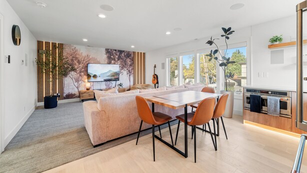 A modern living room with a sofa, dining table, and chairs. There is a TV mounted on the wall, a guitar, and a plant.