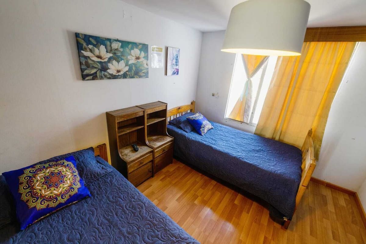 Une petite pièce bien éclairée, avec deux lits simples, une table de chevet en bois, une lampe et une fenêtre avec des rideaux.