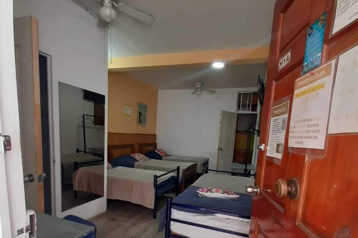 Une chambre avec deux lits simples, un bureau et une chaise. Il y a un ventilateur de plafond et une porte avec une pancarte.