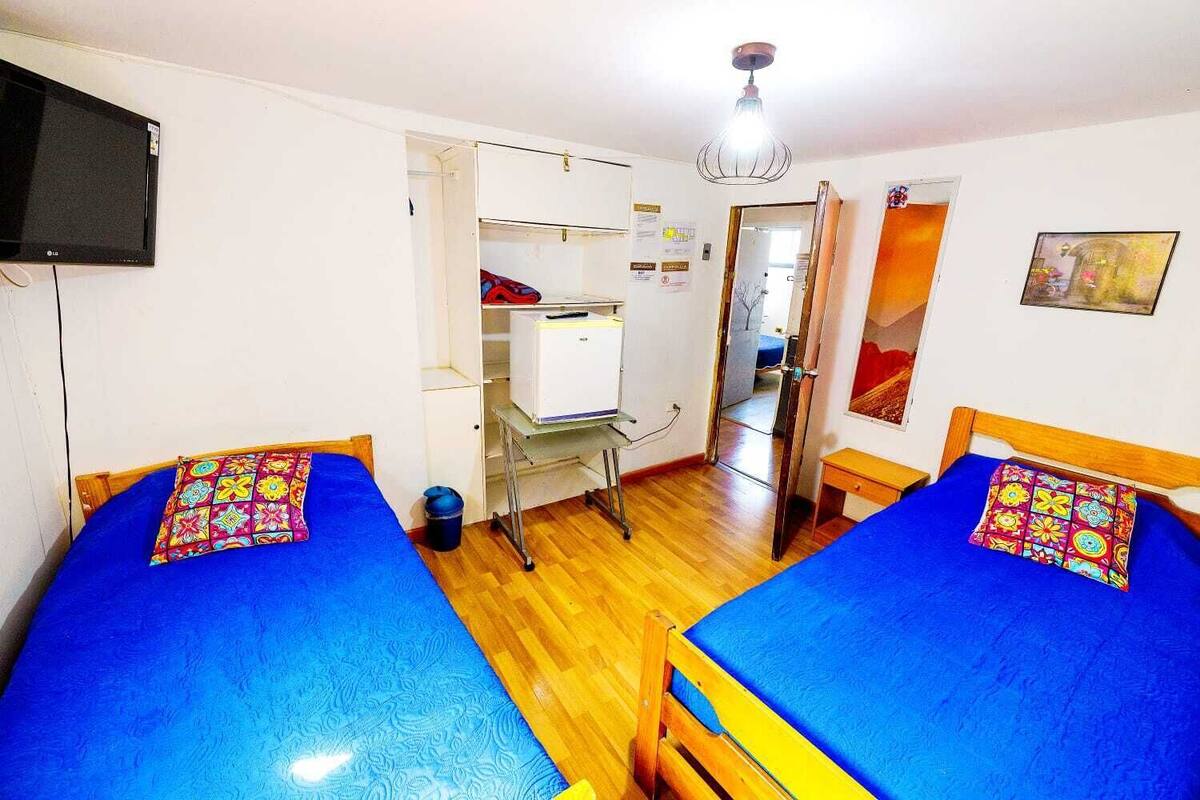 Une chambre avec deux lits, un bureau, une armoire, une télévision et une porte donnant sur une autre pièce.