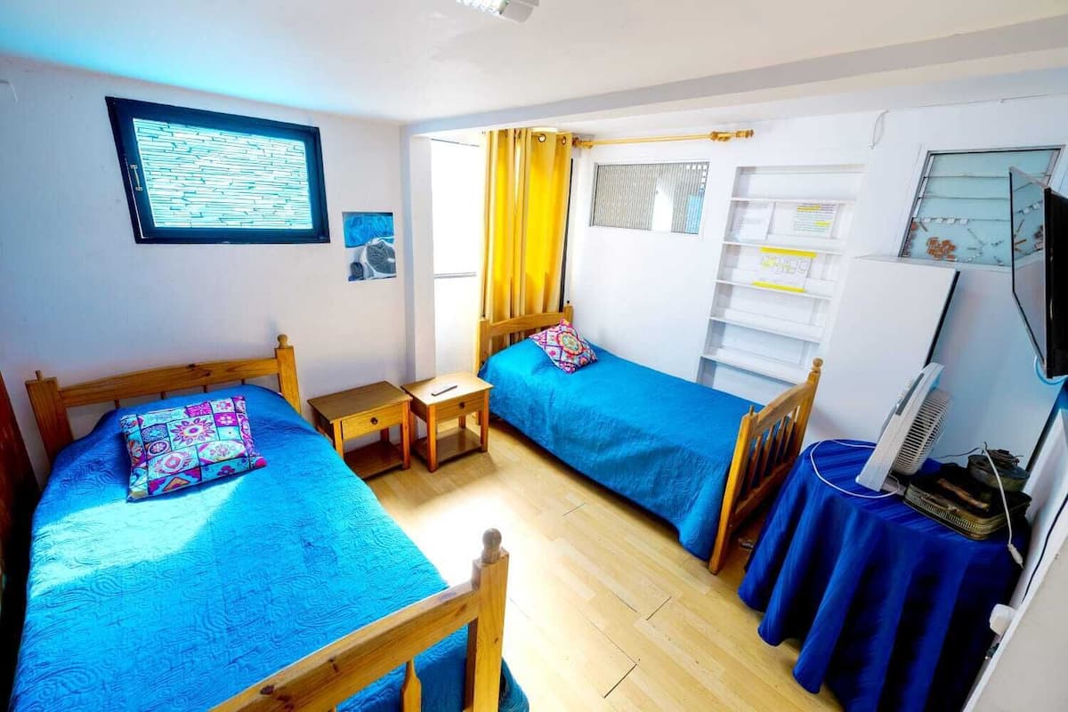 Une chambre avec deux lits simples, une petite table, un téléviseur et une fenêtre avec des rideaux.