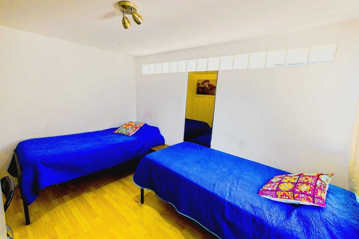 Une chambre avec deux lits simples, chacun recouvert d’une couvre-lit bleue et agrémenté de coussins colorés. La chambre a un plancher en bois et des murs blancs.