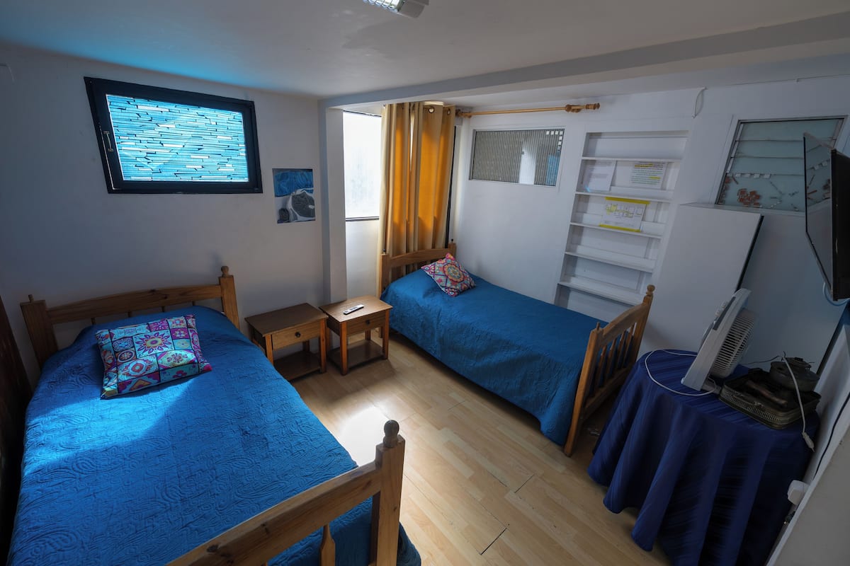 Une pièce avec deux lits, une petite table, une fenêtre avec des rideaux, une étagère avec des papiers et un ventilateur posé sur une table.