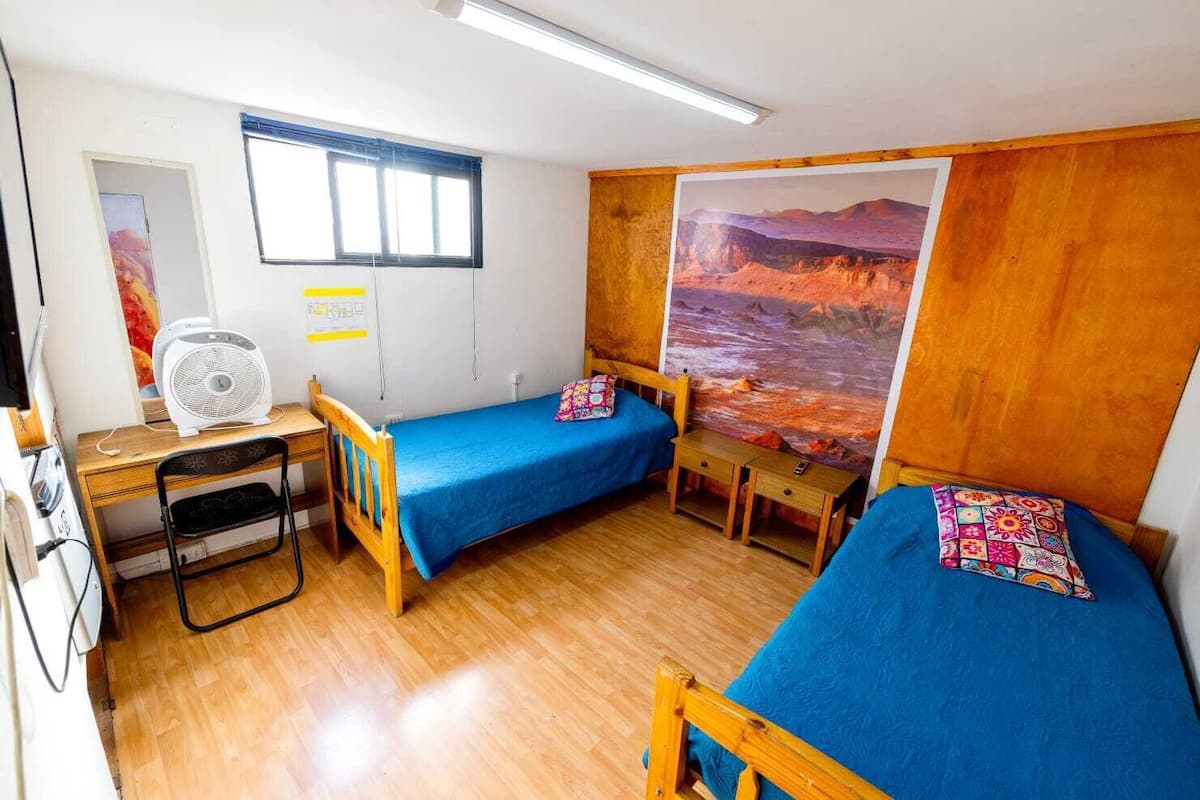 Une chambre de taille modeste, équipée de deux lits simples, d’un bureau, d’une chaise, d’un ventilateur sur pied et d’une grande fresque murale.