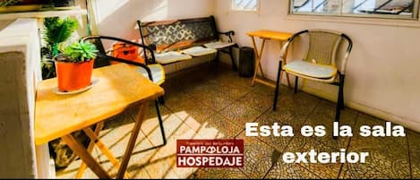 Una habitación pequeña e iluminada por el sol, con un banco, una mesa y una silla. Hay plantas en macetas y una caja eléctrica en la pared.