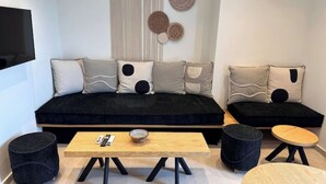 A modern living room with a black sofa, two round ottomans, a wooden coffee table, and a TV mounted on the wall. - Aristotle Suites, Althea

 (Adamantas)