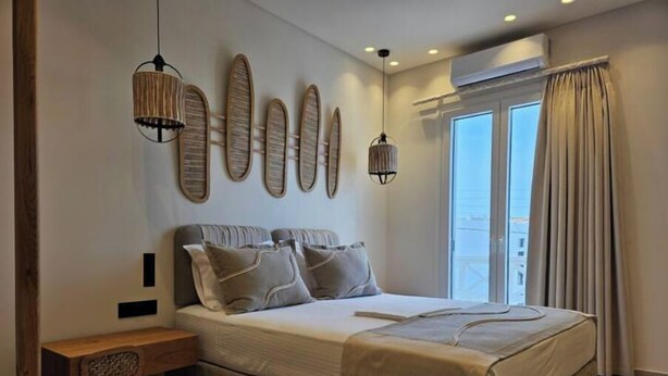 A modern bedroom with a large bed, a bedside table, and a bench. There are wall-mounted lights and a window with curtains. - Aristotle Suites, Althea

 (Adamantas)
