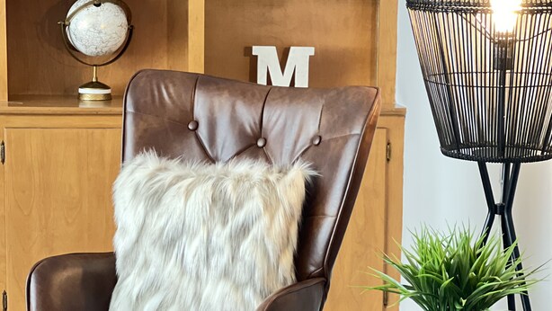 A brown leather chair with a fluffy white pillow, a wooden table with a plant, and a lamp with a woven shade. - Charming 3-bedroom house in peaceful neighborhood  (Bloomington)