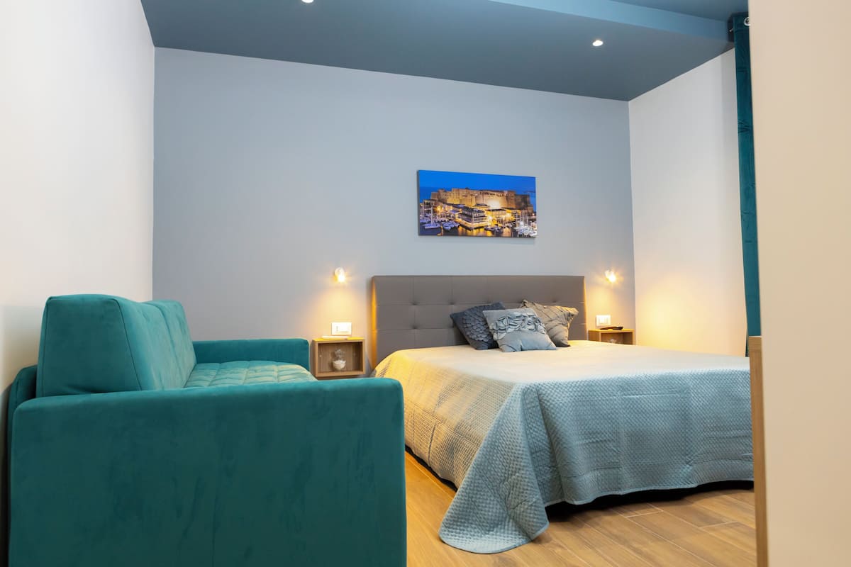 A bedroom with a bed, a teal armchair, a nightstand, and a wall-mounted picture.
