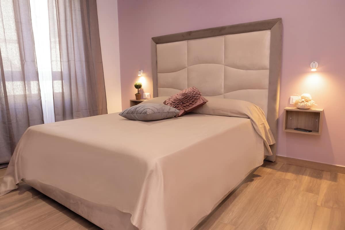 A neatly made bed with a headboard, bedside tables with lamps, and a window with sheer curtains.