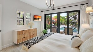 A bedroom with a large bed, a TV, a wooden cabinet, and a view of an outdoor pool area.