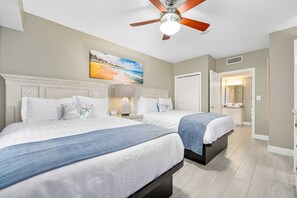 Ein Hotelzimmer mit zwei Betten, einem Deckenventilator, einem Gemälde und einem durch eine offene Tür einsehbaren Badezimmer.
