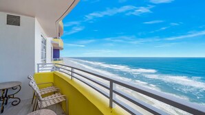 A balcony with a yellow railing, a small table, and a chair, overlooking a beach with waves.
