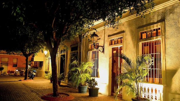 A nighttime street scene with a building featuring a tiled entrance, potted plants, and a motorcycle parked outside. - Honky Tonk Colonial (Santo Domingo)
