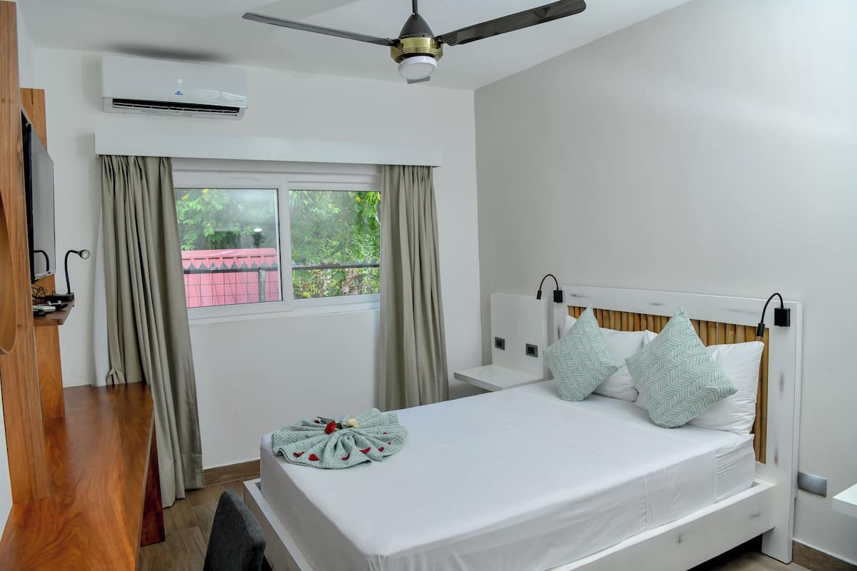 A bedroom with a bed, a bedside table, a ceiling fan, an air conditioning unit, a window with curtains, and a view of greenery outside.