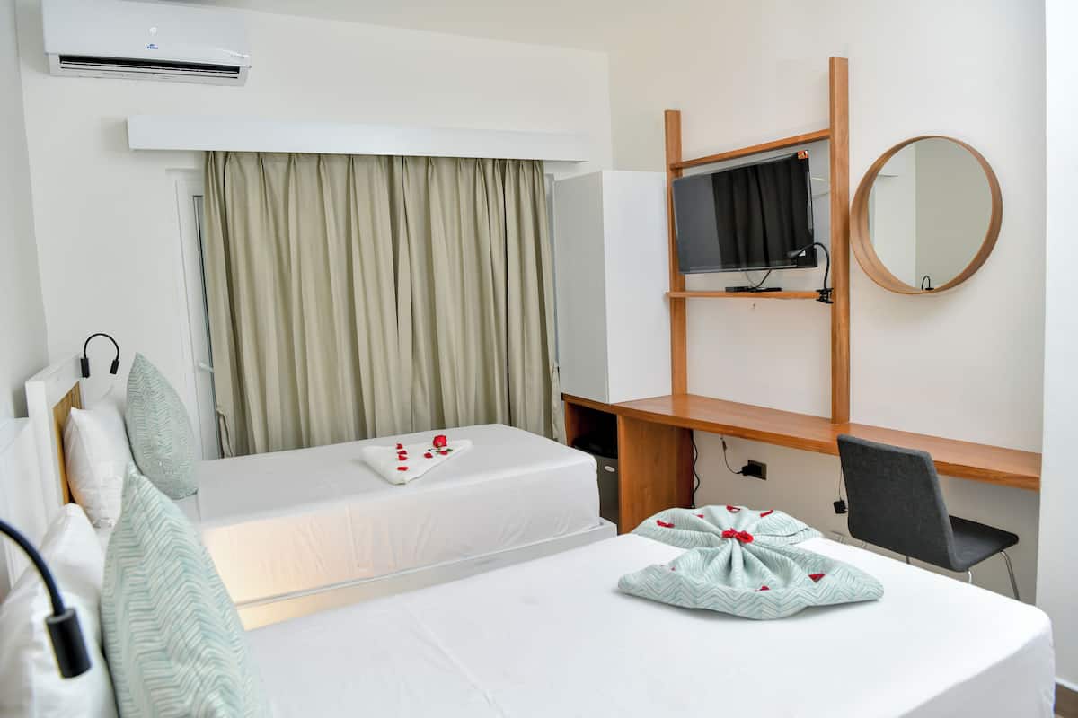 A hotel room with a bed, a desk with a computer, a chair, a round mirror, and an air conditioning unit.