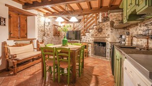 Een rustieke keuken met stenen muren, houten balken, een eettafel met stoelen, een open haard en een groen kastje.