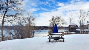 A snow-covered picnic area with a bench and a blue umbrella, overlooking a body of water and buildings in the background.