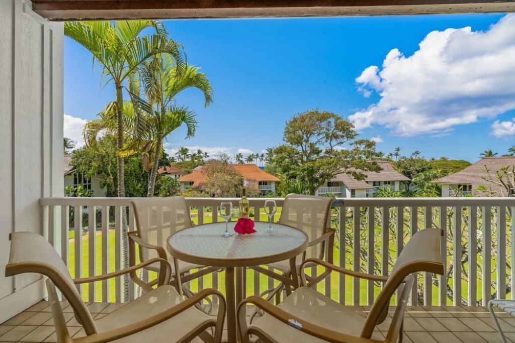 Un balcon avec une table ronde et des chaises, donnant sur un paysage tropical avec des palmiers et des maisons.