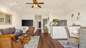 A spacious living room with a sofa, chairs, and a television. A kitchen area with a refrigerator and a dining table is visible in the background. - Kiahuna Paradise condo with A/C (Koloa)