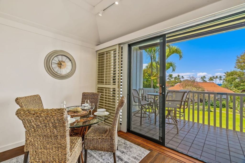 A dining area with wicker chairs and a table set for two, a large window with blinds, and a view of a garden and palm trees.