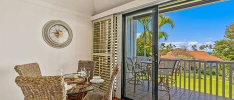 A dining area with wicker chairs and a table set for two, a large window with blinds, and a view of a garden and palm trees.