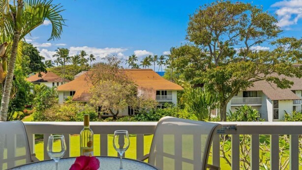 A table with two wine glasses and a bottle, set against a backdrop of a tropical landscape with palm trees and houses. - Kiahuna Paradise condo with A/C (Koloa)