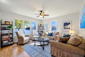 A living room with a ceiling fan, a glass coffee table, a beige sofa, a wicker chair, a bookshelf, and large windows with a view of trees and a building. - Lakefront Lake Anna - Views, Boat Dock, Games (Mineral)