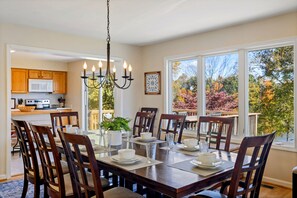 A dining area with a large table set for six, surrounded by wooden chairs, a chandelier, and a view of autumn foliage through large windows.