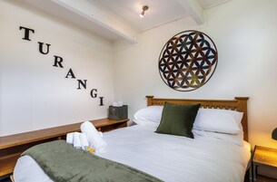 Uma cama bem arrumada com lençóis brancos, um cobertor verde e um travesseiro. Há uma cabeceira de madeira, uma mesa de cabeceira com uma luminária e uma decoração na parede com a palavra "TURANGI".
