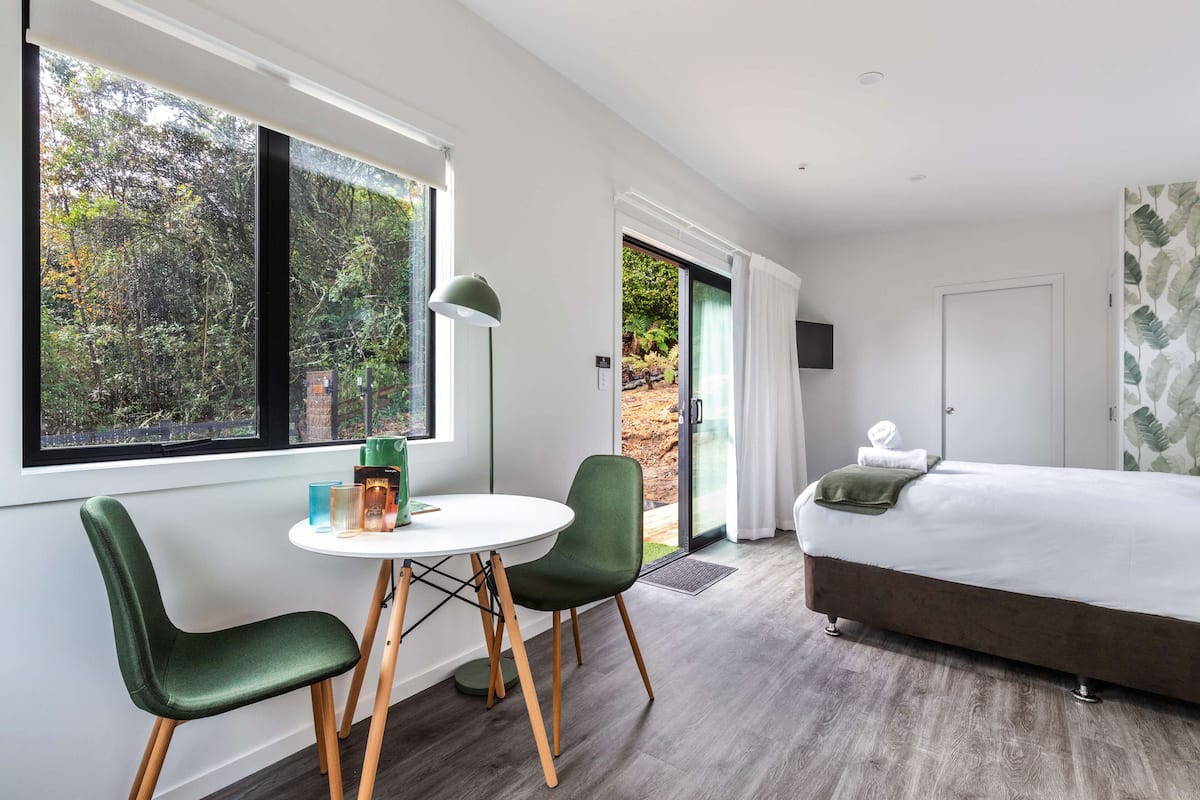 A modern bedroom with a large bed, a dining table with chairs, and a view of a forest through the window.