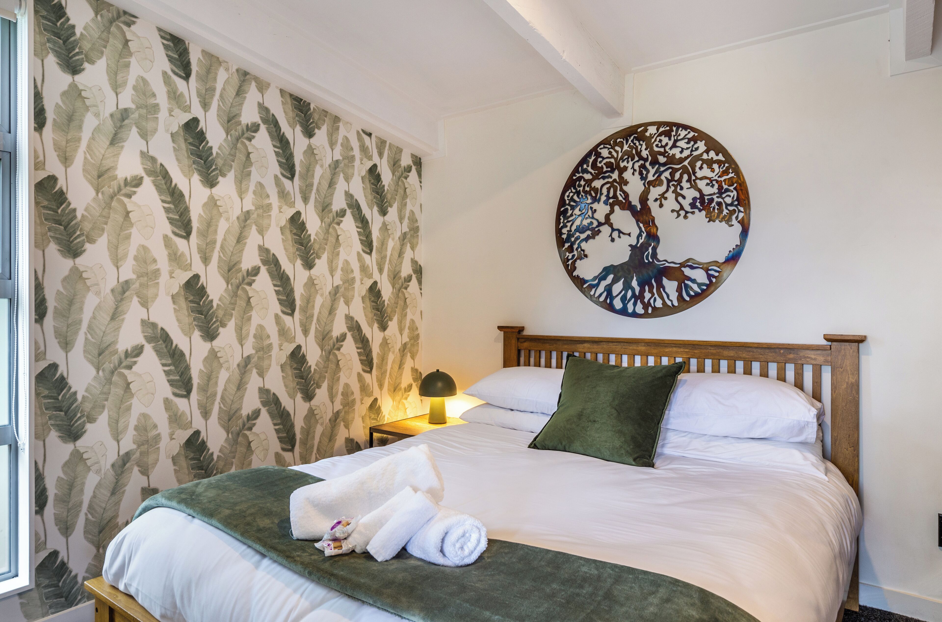 A neatly made bed with white linens, a green throw blanket, and rolled towels. A round wall art depicting a tree is above the bed.