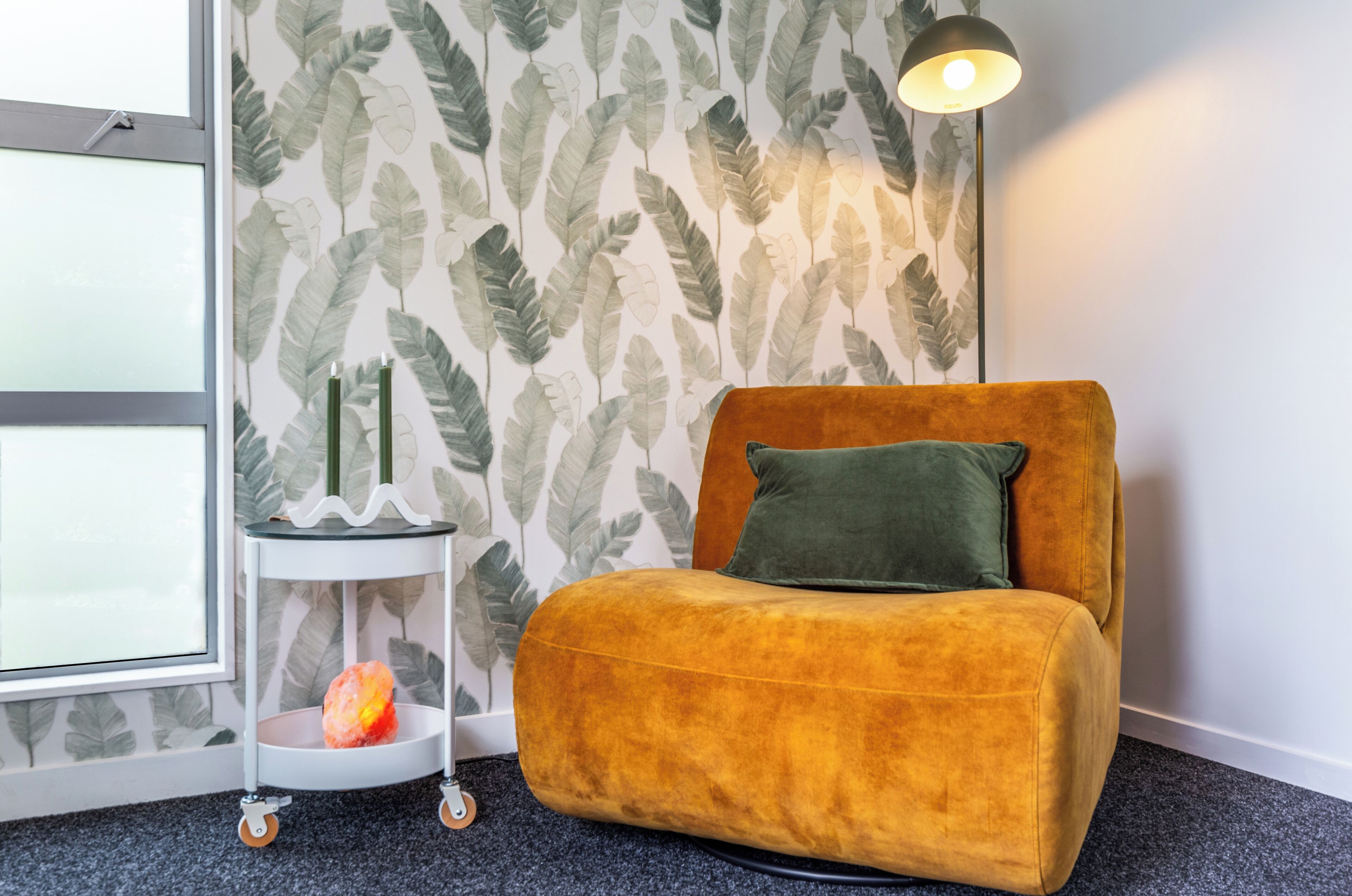 Ein Zimmer mit einem senffarbenen Sofa, einem grünen Kissen, einem kleinen Beistelltisch mit einer Kerze und einer Wand mit einem Blattmuster.