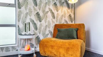 Ein Zimmer mit einem senffarbenen Sofa, einem grünen Kissen, einem kleinen Beistelltisch mit einer Kerze und einer Wand mit einem Blattmuster.