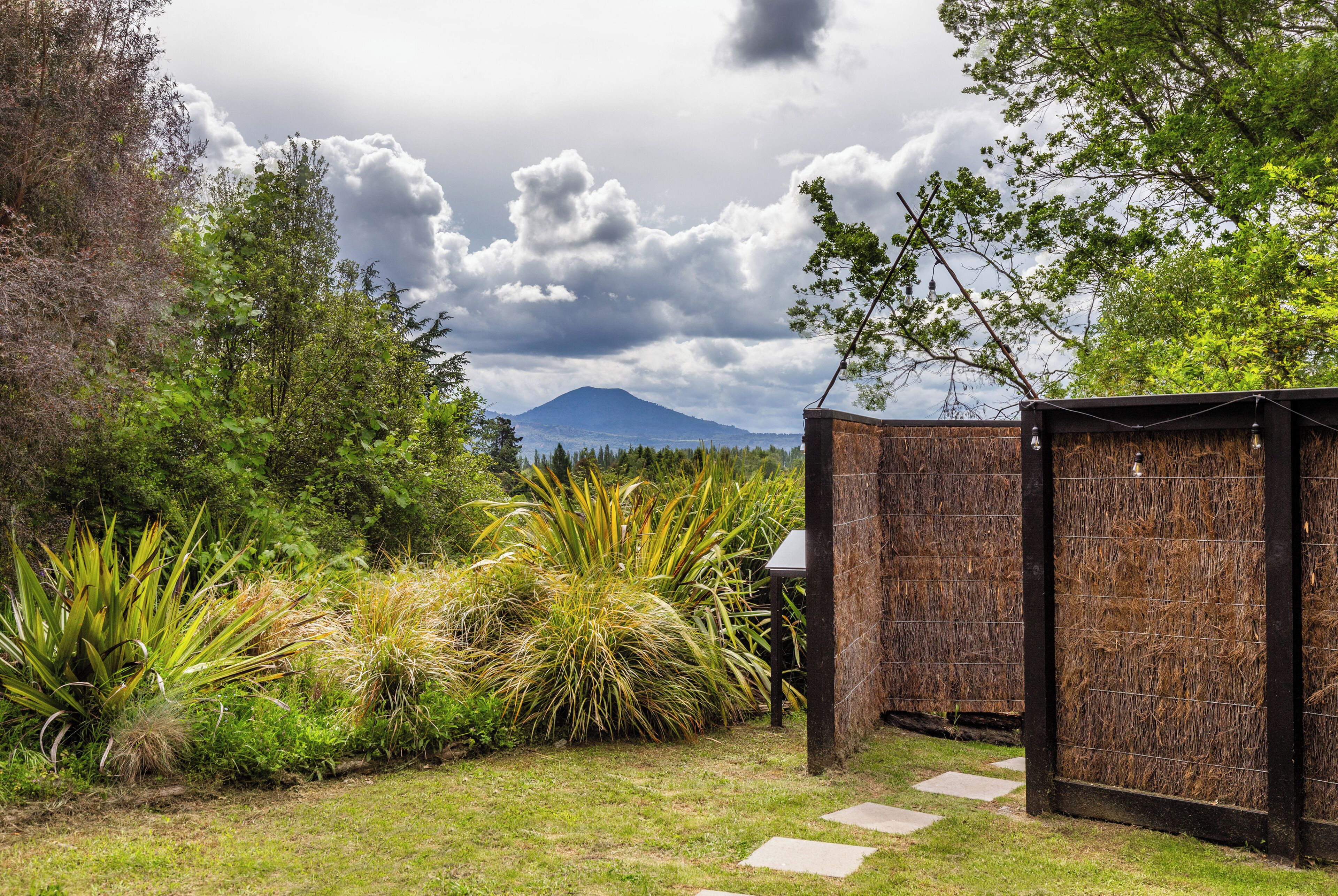 Un giardino con un sentiero che conduce a un cancello di legno, una montagna sullo sfondo e un cielo nuvoloso.