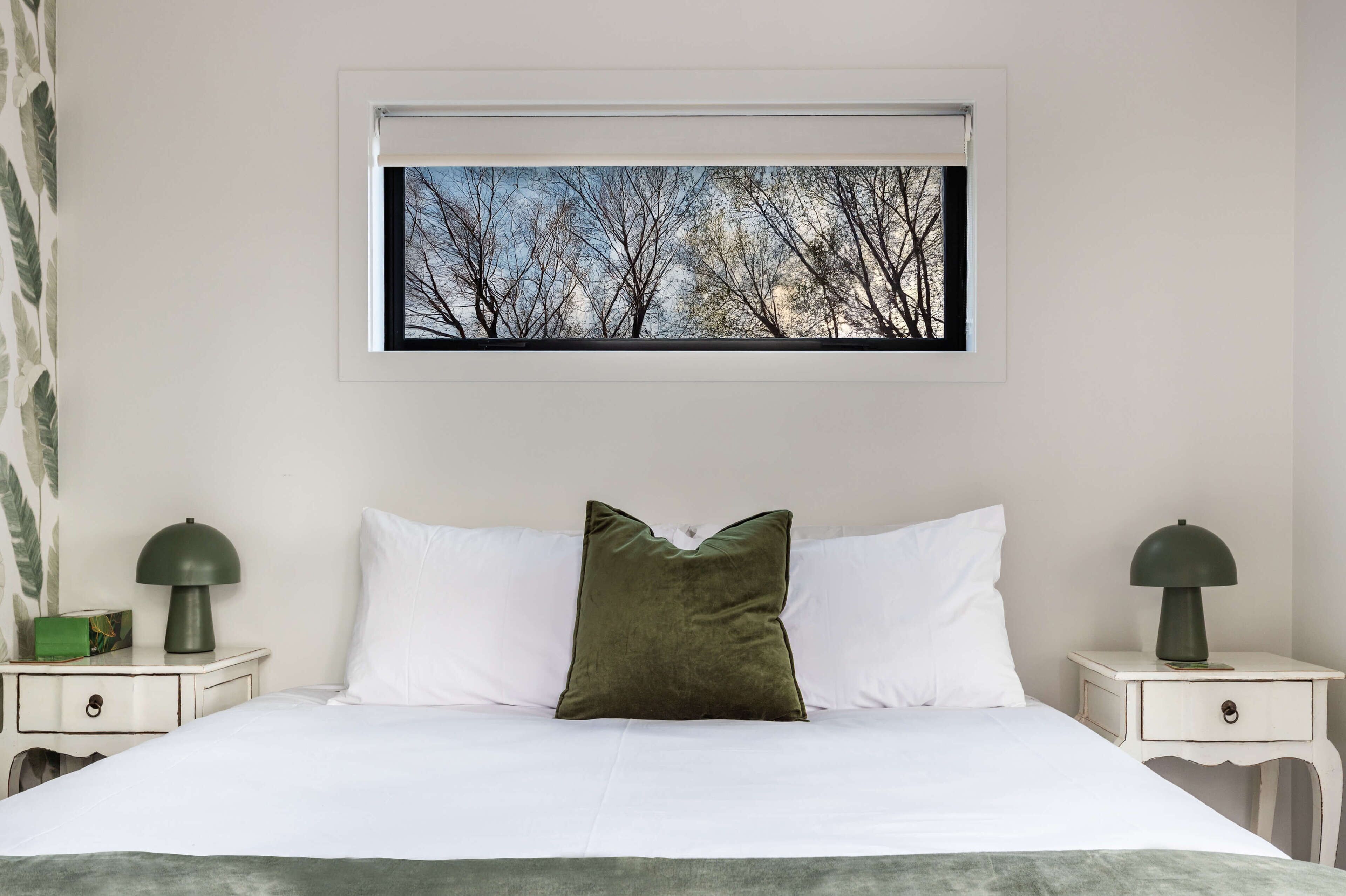 A neatly made bed with white linens and a single green pillow, flanked by two bedside tables with lamps, one green and one black. A framed picture of trees is hung above the bed.