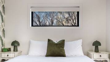 A neatly made bed with white linens and a single green pillow, flanked by two bedside tables with lamps, one green and one black. A framed picture of trees is hung above the bed.